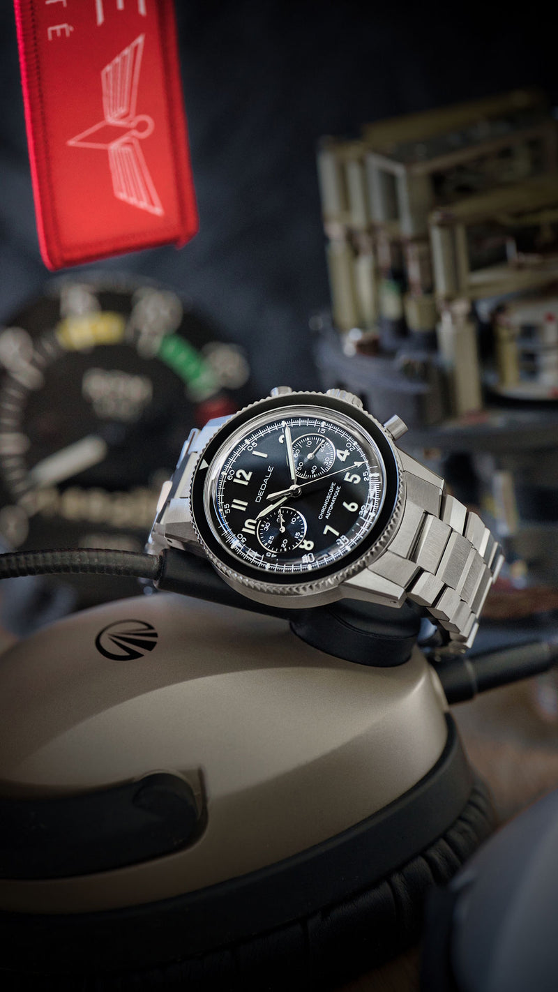 Montre française aéronautique DÉDALE WP-01 NOVEMBER, cadran noir et bracelet en acier inoxydable 316L WingSteel, posée sur un casque de pilote, devant des instruments d'avion.
